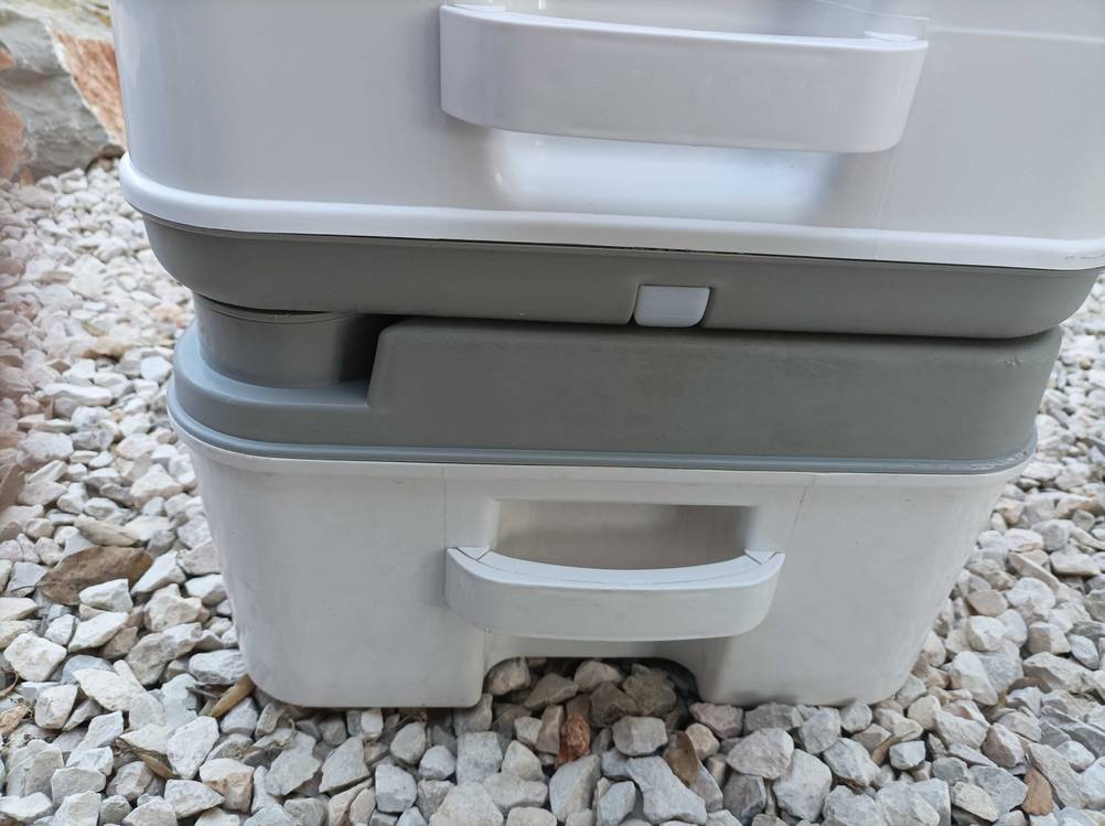 Getestete Campingtoilette im grau-weißen Design steht zusammengebaut, mit Fokus auf die integrierten Handgriffe, auf Schottersteinen.