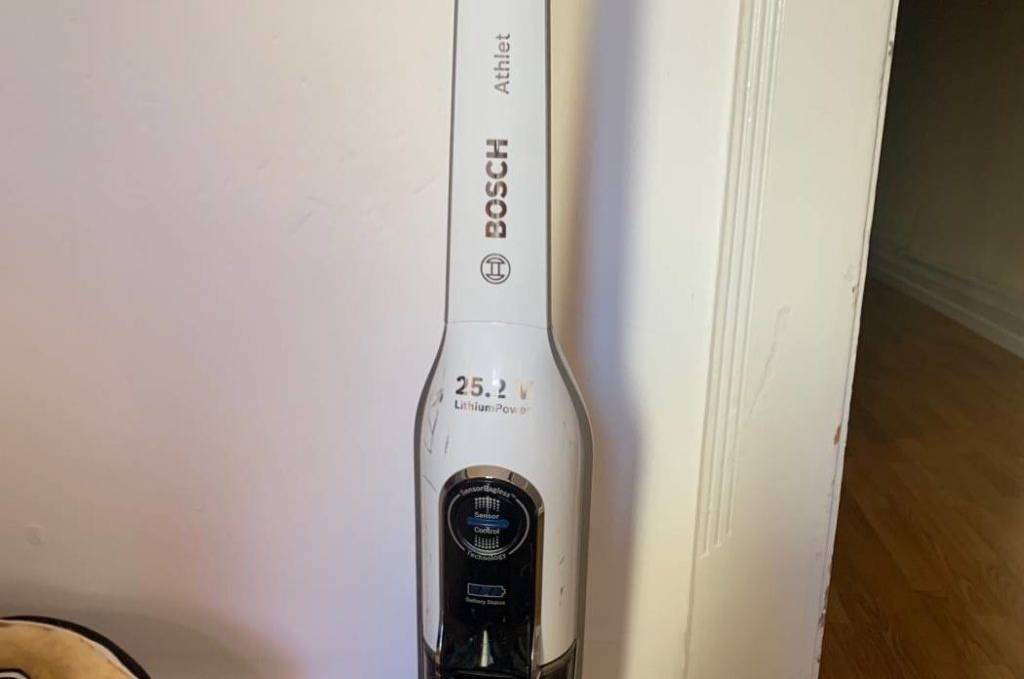 Bosch-Akkusauger-Test: beutelloser Bosch BBH32551 in weißem Design mit transparentem Auffangbox und 25.2 Volt Leistungskapazität steht an einer weißen Wand.
