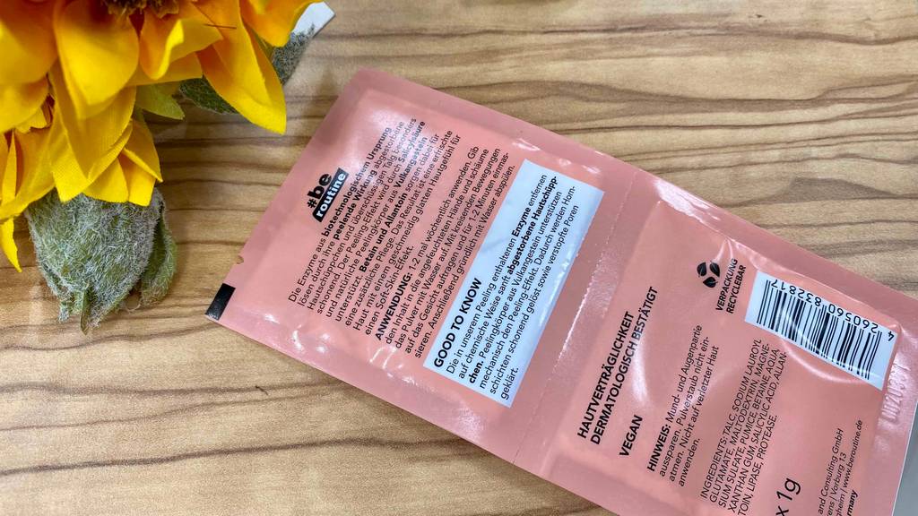 Verpackung eines Enzympeelings liegt mit der Rückseite nach oben auf einem Holztisch mit Blumen im Hintergrund.