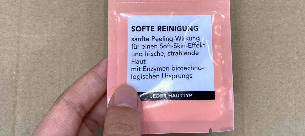 Hand hält den rosa Beutel mit dem getesteten Enzympeeling-Pulver vor einem beigefarbenen Hintergrund.