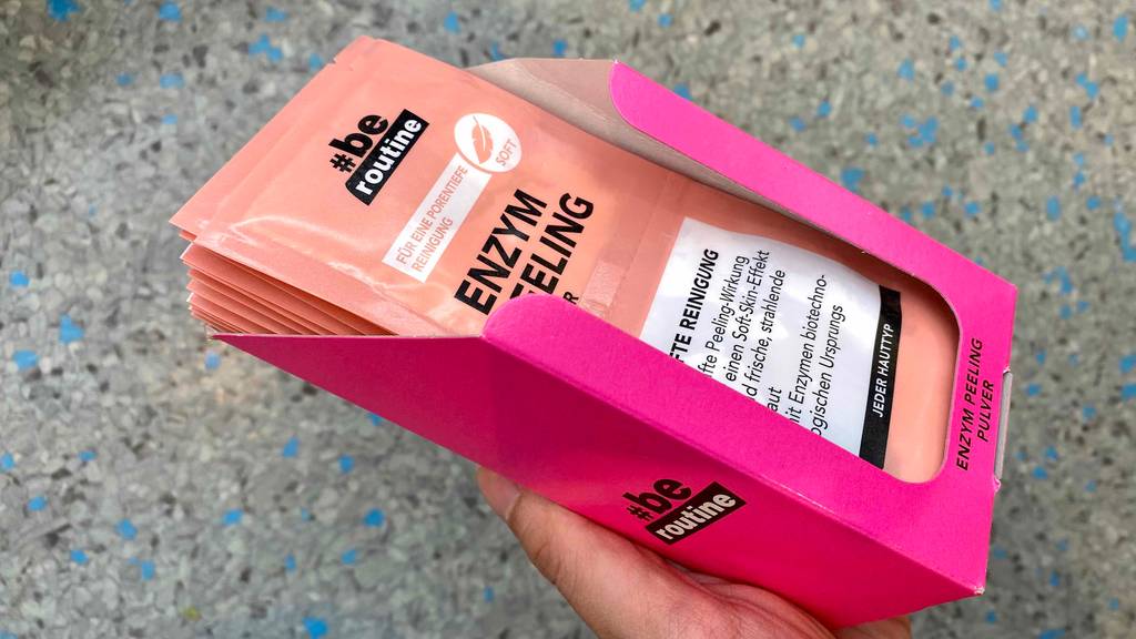 Getestetes Enzympeeling-Pulver: Hand hält eine rosafarbene Verkaufsschachtel mit mehreren Zweierkonfektionen vor einem grauen Hintergrund.