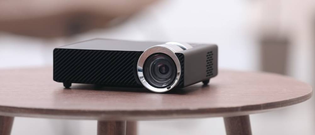 Beamer-unter-100-Euro-Test: Ein schwarzer kleiner Beamer steht auf einem runden Holztisch.