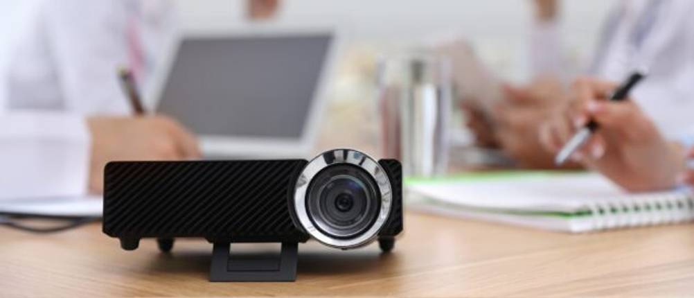 Beamer-unter-100-Euro-Test: Ein schwarzer Mini-Beamer steht in einem Meeting auf einem Holztisch.