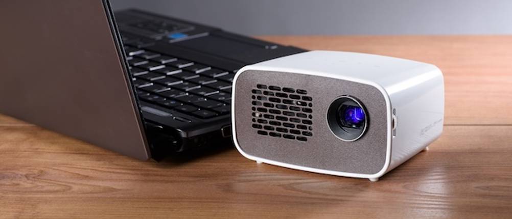 Beamer unter 100 Euro getestet: Ein weißer Mini-Beamer steht auf einem Holztisch neben einem Laptop.