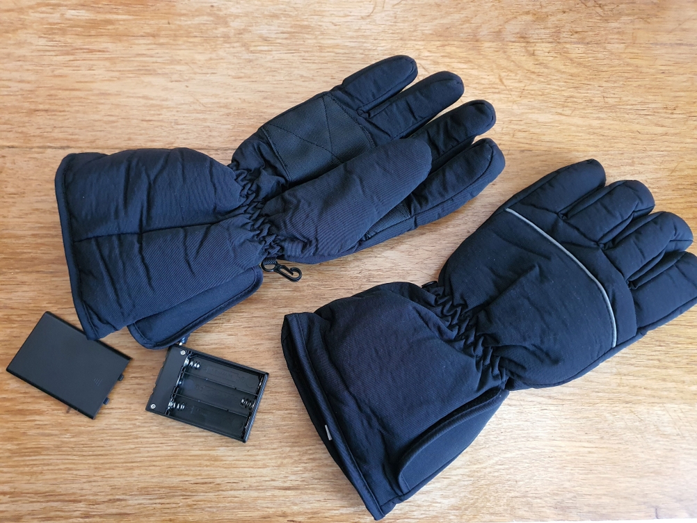 Beheizbare Handschuhe und der dazugehörige Akku liegen auf einem Holzuntergrund.
