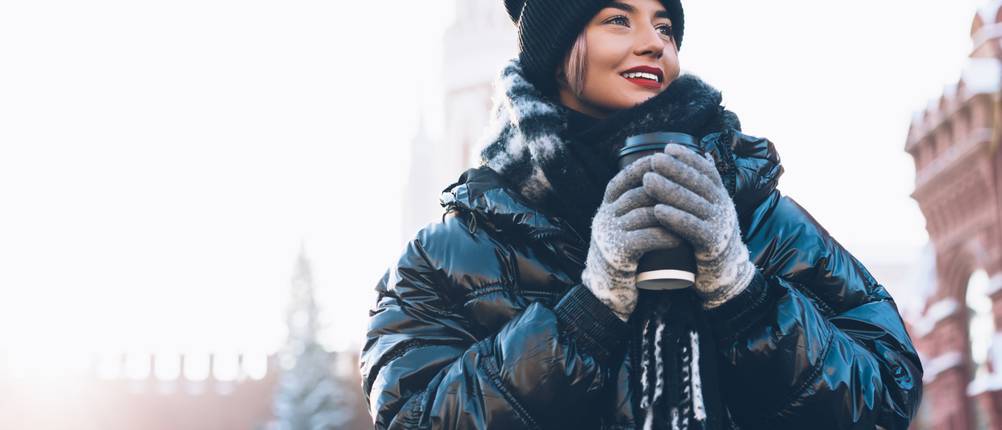 Beheizbare Jacke für Damen im Test: Frau mit einer dick gefütterten Wintersteppjacke, die ein heißes Getränk in ihren behandschuhten Händen hält