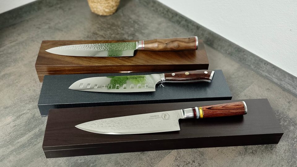 Drei Damastmesser verschiedener Marken liegen auf ihren Verpackungen. Die Messer sind vollständig sichtbar und nebeneinander platziert.