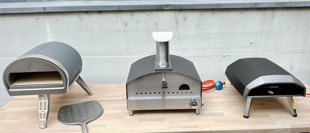 Drei Gas-Pizzaöfen stehen nebeneinander auf einem Tisch: Gozney, Heidenfeld und Ooni. Zubehör wie Schaufeln ist ebenfalls sichtbar.