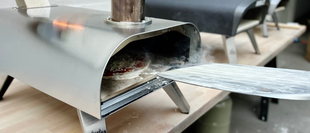 Der Big-Horn-Outdoors-SRPG18003-Pizzaofen mit eingelegter Pizza steht in Betrieb auf einer Werkbank. Rauch steigt aus der Öffnung.