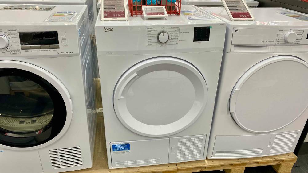 Ein getesteter Beko-Trockner zwischen anderen Geräten auf einem Holzsockel in einer Verkaufsfläche inklusive Preisschilder.