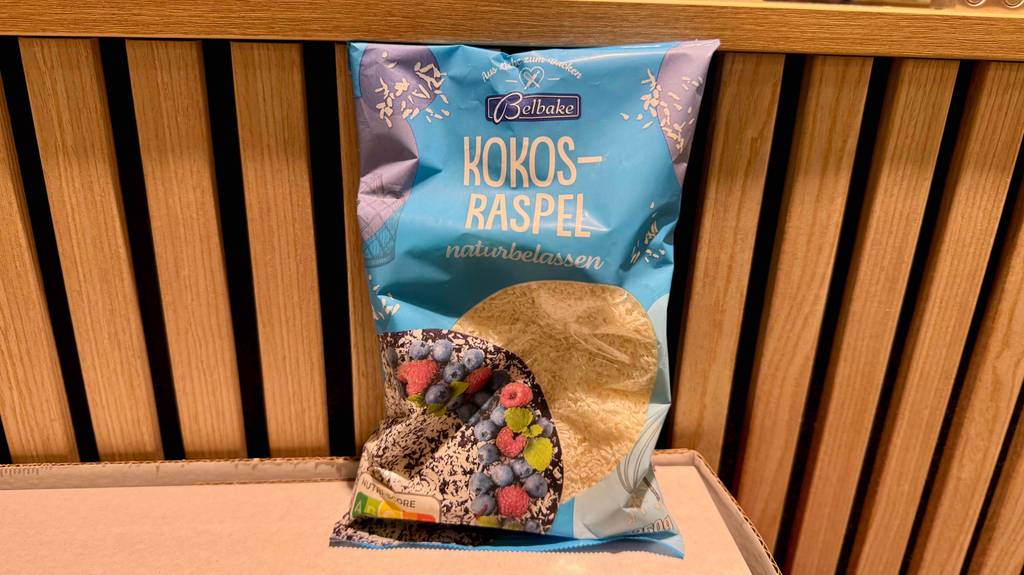 Eine Packung Kokosraspeln steht zum testen vor einem Holzregal.