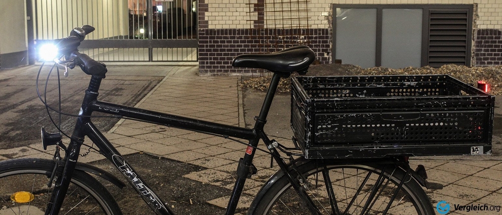 Fahrradbeleuchtung im Test: Seitenaufnahme eines beleuchteten Fahrrads vor einem Industriegebäude, auf dem Gepäckträger befindet sich eine Plastikkiste