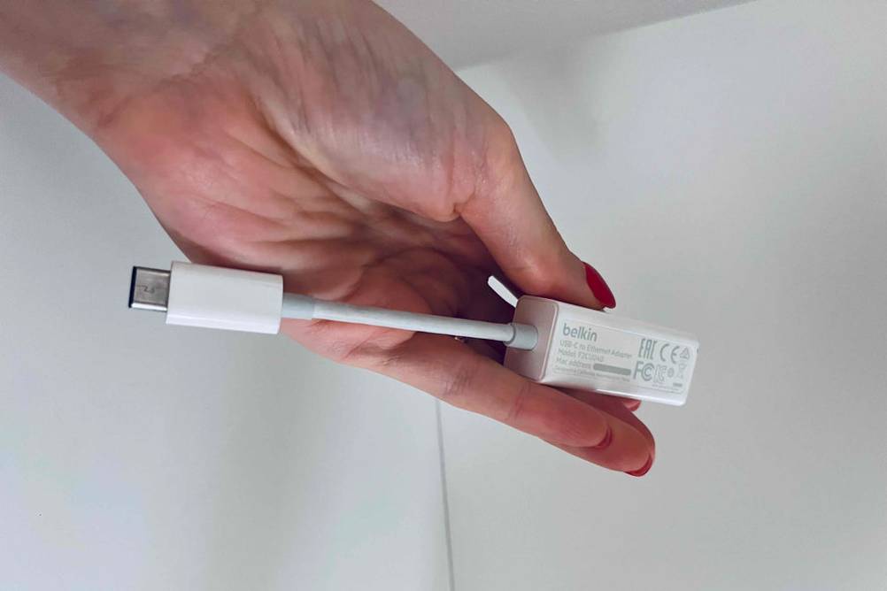 Hand hält einen getesteten Netzwerkadapter von Belkin vor weißem Regal.