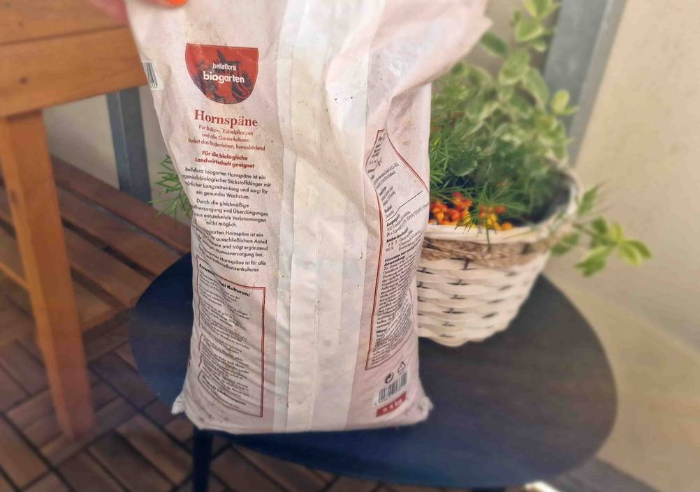 Bellaflora Biogarten Hornspäne im Test befinden sich in einem weißen 2,5-kg-Kunststoffsack vor einer Pflanzenkomposition auf einem schwarzen Hocker.