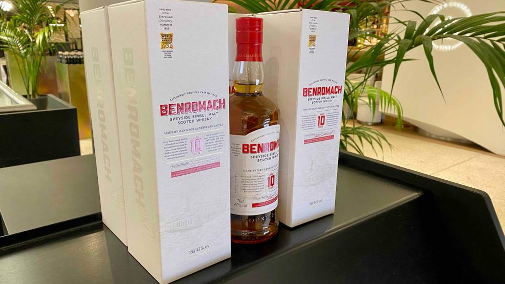 Eine Flasche getesteten Single-Malt-Scotch-Whiskys steht zwischen zwei Originalverpackungen auf einer schwaren Fläche im Hintergrund Pflanzen.