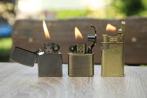 Benzinfeuerzeuge getestet: Drei Benzinfeuerzeuge stehen mit brennender Flamme nebeneinander auf einem Holztisch.