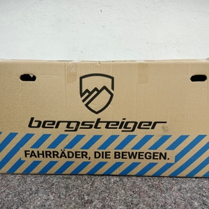Das Foto zeigt den Versandkarton des Bergsteiger Windsor mit Herstellerlogo, Schriftzug und Tragegriffen auf einer Schutzdecke.