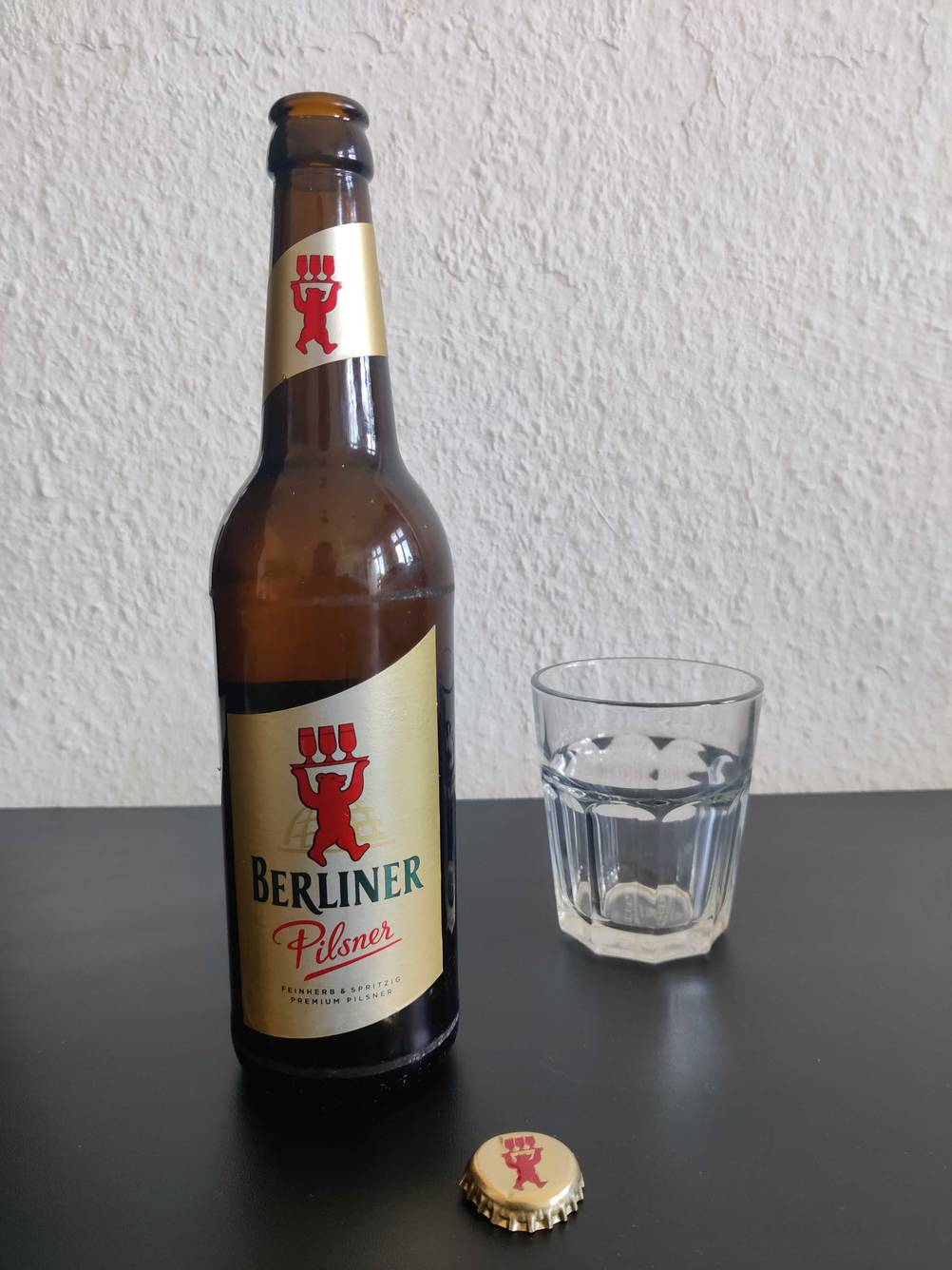 Bier-Test: Eine Flasche neben einem Kronkorken und einem Glas auf einer dunklen Fläche.