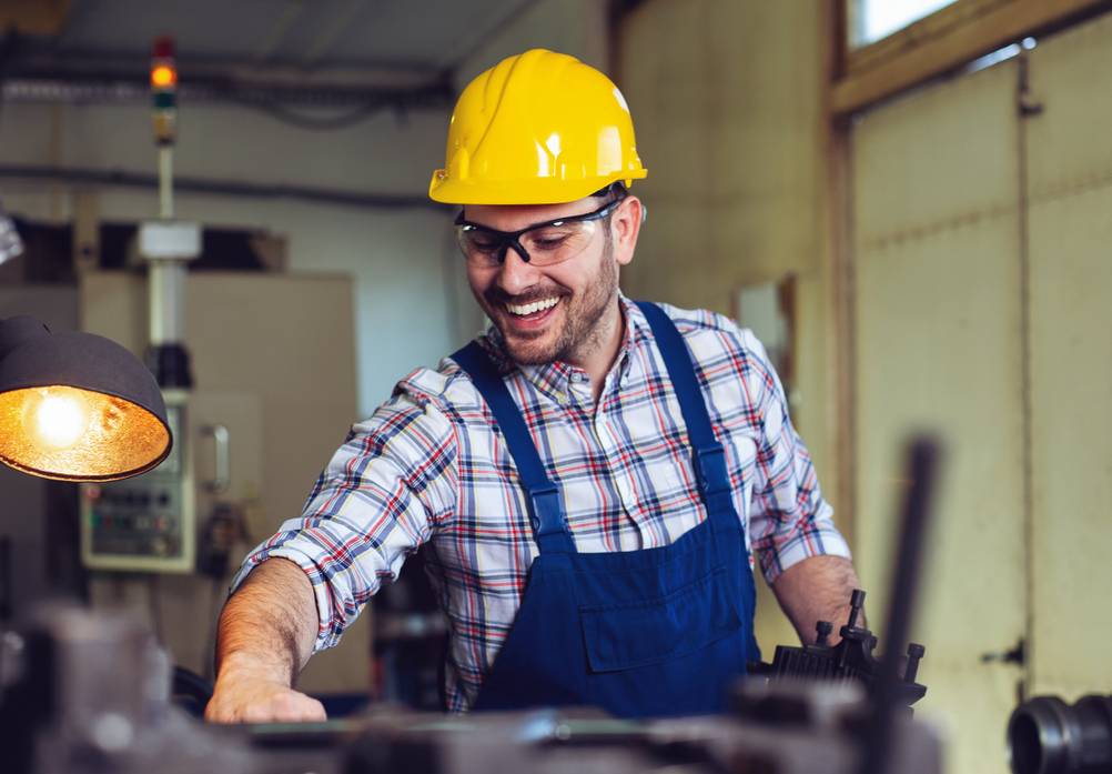 Bernardo-Drehmaschine im Test: Ein Mann mit gelbem Helm und blauer Latzhose steht in einer Werkstatt vor einer Maschine und lächelt.