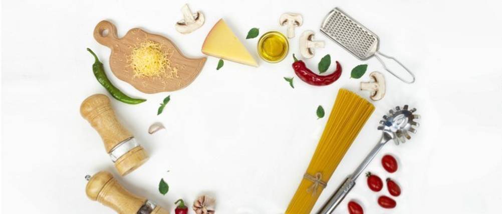 Bertolli-Olivenöl-Test: Auf einem hellen Untergrund befinden sich eine kleine Schale mit Olivenöl, Spaghetti, Pfeffer- und Salzstreuer, ein Schneidebrett mit Parmesan, eine Reibe, aufgeschnittene Champignons, Tomaten, Basilikum, Knoblauch und Pfefferoni. 