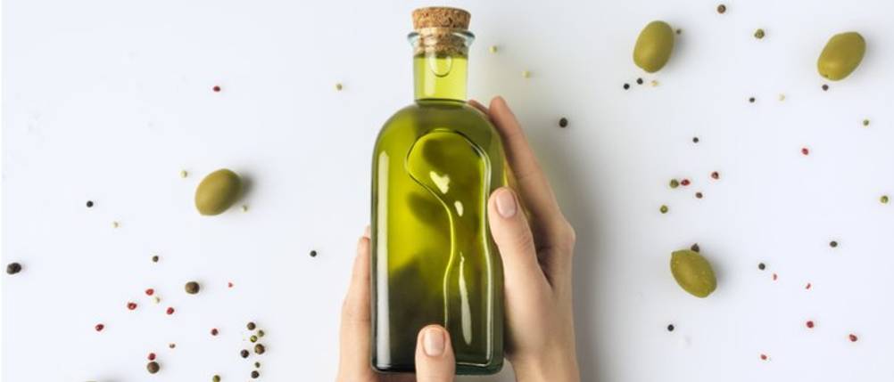 Bertolli-Olivenöl-Test: Zwei Hände umfassen eine Glasflasche mit Olivenöl. Daneben liegen grüne Oliven und Pfefferkörner. 