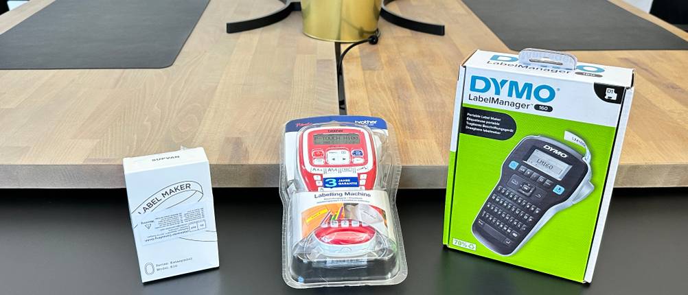Drei verpackte Beschriftungsgeräte von Brother, Dymo und Supvan liegen nebeneinander auf einem schwarzen Tisch.