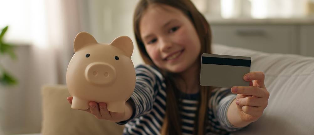 Ein kleines Mädchen hält ein Sparschwein und eine Bankkarte nach oben.