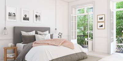 Ein gemachtes Bett in einem aufgeräumten Schlafzimmer. Im Hintergrund ist ein geöffnetes Fenster zu sehen. Über dem Bett hängen Bilder