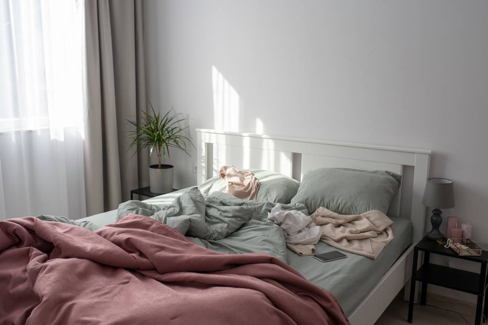 Ein Doppelbett mit zerknitterter Bettdecke und chaotisch herumliegenden Textilien.