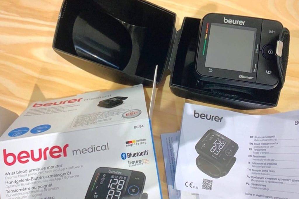Handgelenk-Blutdruckmessgerät im Test: Ansicht von oben der Originalverpackung eines Beurer Medical BC 54 Modells, sowie schwarzes Messgerät in Aufbewahrungsbox und Bedienungsanleitung auf hellem Holzuntergrund.