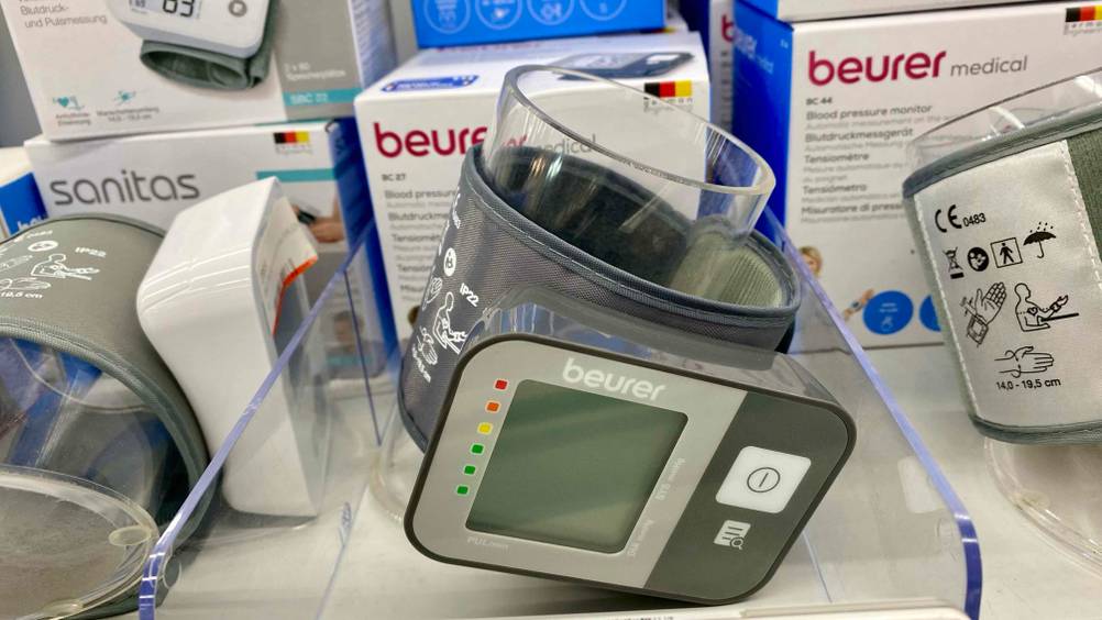 Getestet: Ein Beurer-Blutdruckmessgerät an einer transparenten Rolle befestigt in einem Verkaufsregal zwischen anderen Produkten.