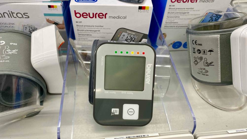 Ein getestetes Beurer-Blutdruckmessgerät auf der Seite liegend von vorne in einem Verkaufsregal zwischen anderen Produkten.