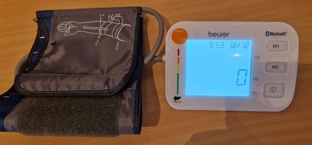 Weißes getestetes Oberarm-Blutdruckmessgerät der Marke Beurer mit großflächiger, beleuchteter LCD-Anzeige befindet sich neben seiner dunklen Armmanschette auf einer Holzfläche.