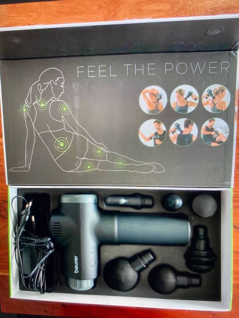Nahaufnahme einer geöffneten Verpackung einer Massagepistole im Test inklusive Bildanleitung und elektrischer Massagepistole mit verschiedenen Aufsätzen.