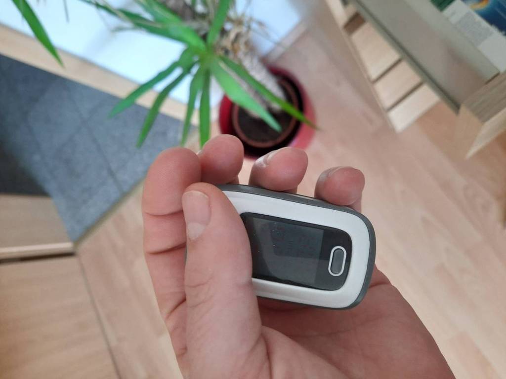 Hand hält einen ausgeschalteten weiß-schwarzen Pulsoximeter im Test, mit Blick auf den Boden eines Wohnraums.