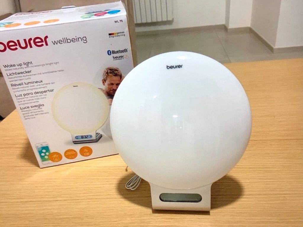 Lichtwecker-Test: Nahaufnahme eines runden, weißen Beurer Wellbeing Lichtweckers. Er steht ausgeschaltet neben seiner Verpackung auf einer Holzfläche.