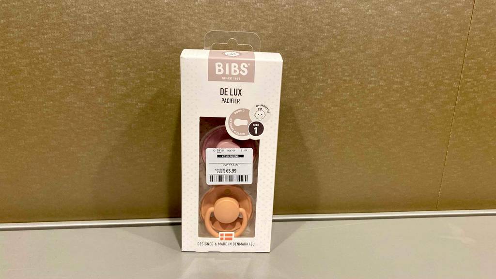Im Test: Bibs-Schnuller in beigefarbener Verpackung mit Sichtfenster, hinter dem zwei Schnuller zu sehen sind, auf einer weißen Oberfläche an einer goldenen Wand lehnend.