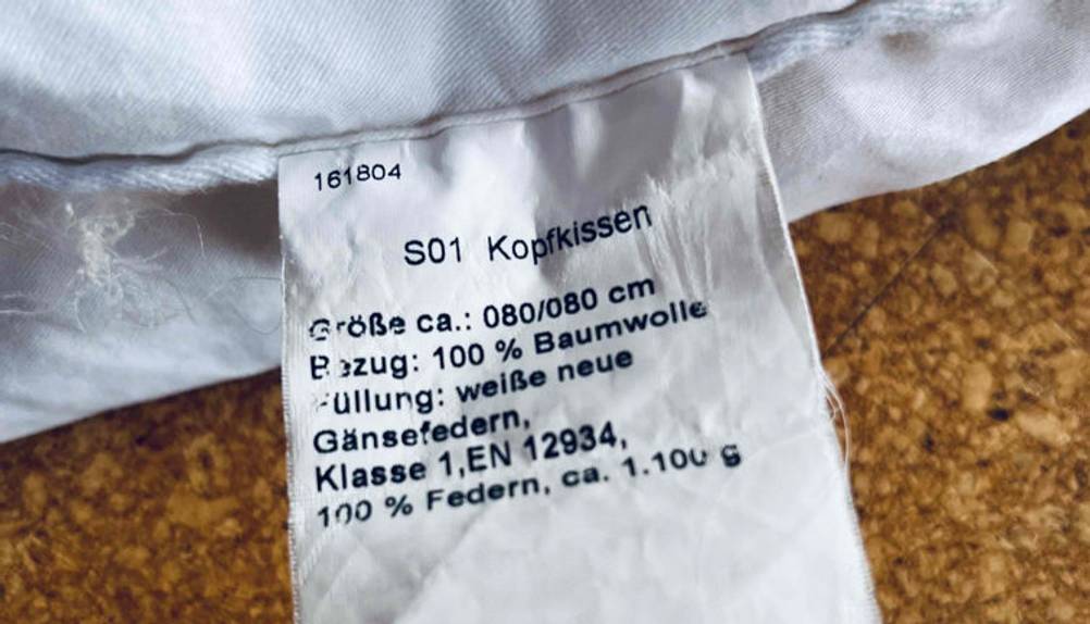 Nahaufnahme des Etiketts eines weißen Baumwoll-Daunenkissens 80×80 cm im Test auf braun gesprenkeltem Untergrund.