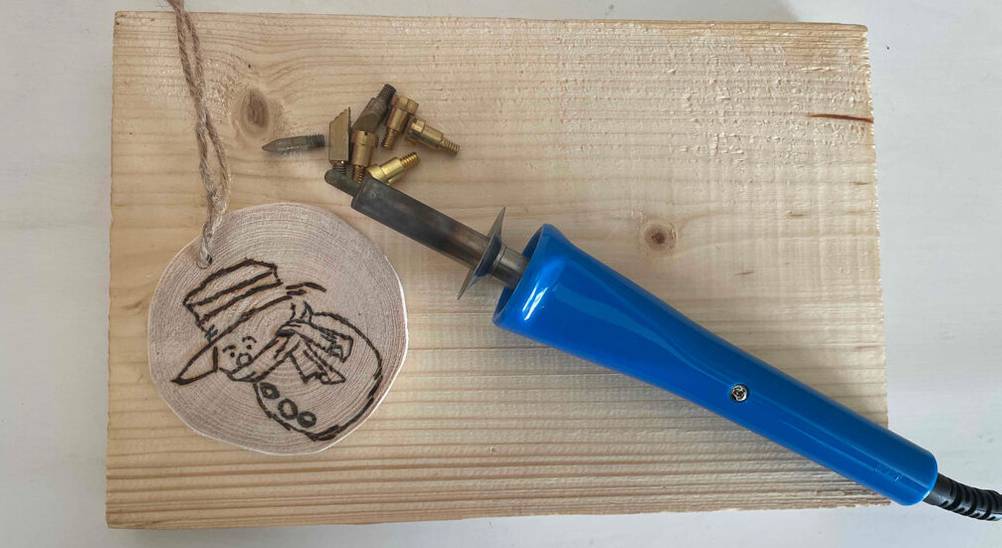 Brandmalkolben im Test: Ansicht von oben auf ein Modell mit blauem Griff, das neben mehreren austauschbaren Spitzen und einer Brandmalerei auf einem Holzbrett liegt.