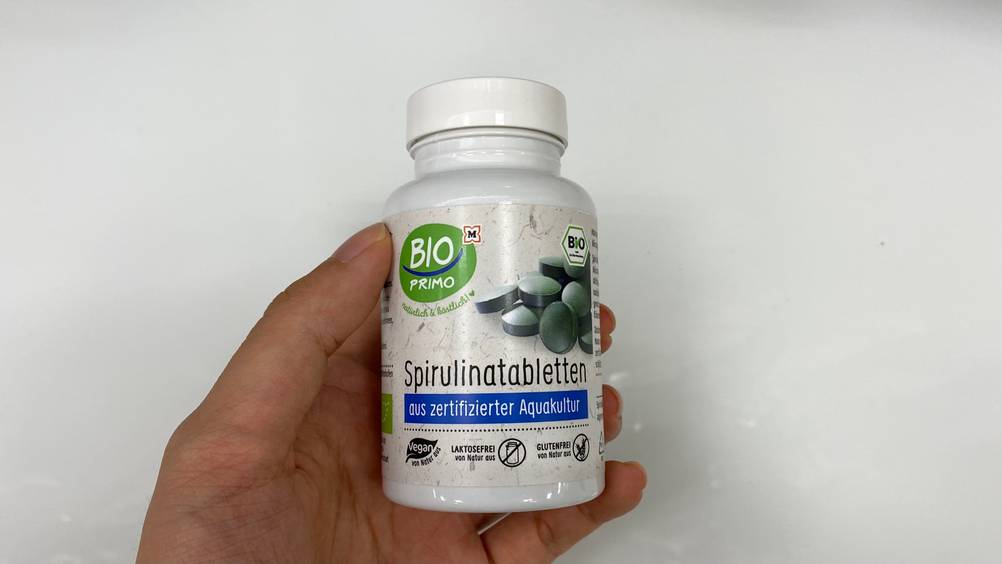 Spirulina-Tabletten-Test: Eine Hand hält eine Dose Spirulina-Tabletten von vorne vor eine helle Fläche.