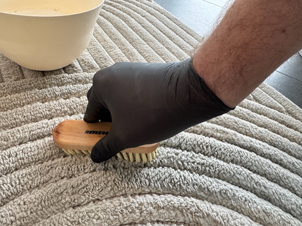 Eine Hand reibt mit einer Bürste den Biohy-Teppich-Shampoo in einen Teppich ein.