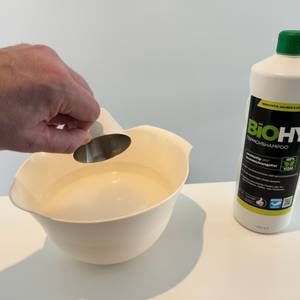 Eine Hand entleert einen Behälter, in dem zuvor der Biohy-Teppich-Shampoo enthalten ist, in eine Schale Wasser.
