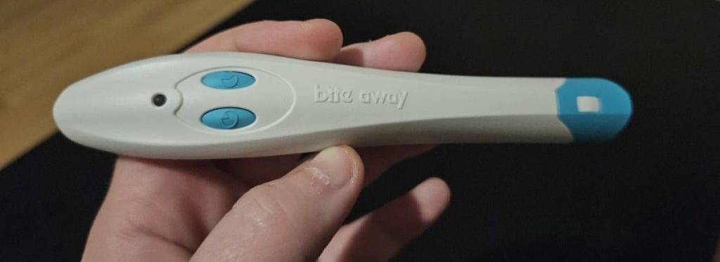 Hand hält weiß-blauen getesteten Stichheiler der Marke bite away vor schwarzem Hintergrund.