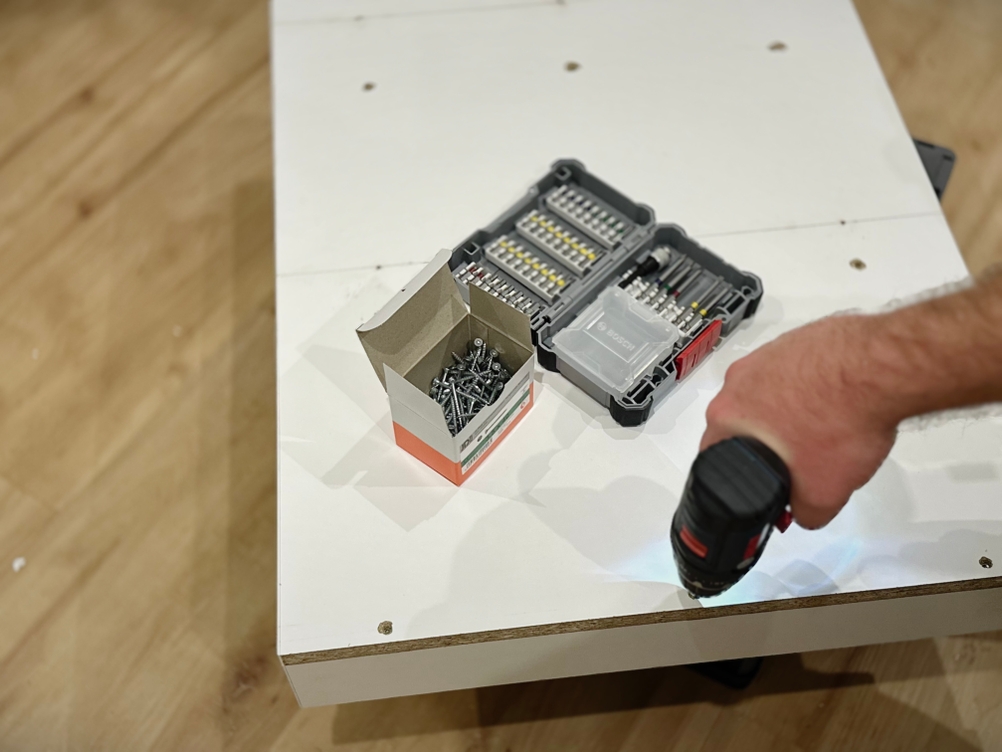 Mit einem Akkuschrauber wird eine Schraube in eine liegende weiße Spanplatte geschraubt.