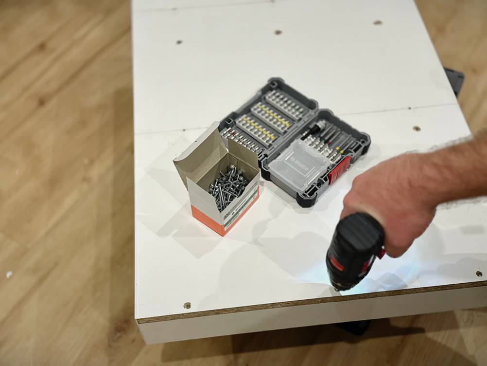 Mit einem Akkuschrauber wird eine Schraube in eine liegende weiße Spanplatte geschraubt.