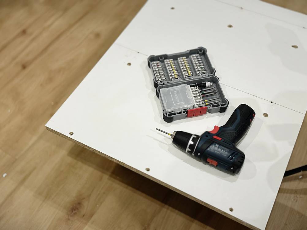 Zu sehen ist das Bitset mit einem Akkuschrauber, diese auf einer weißen Spanplatte liegen.