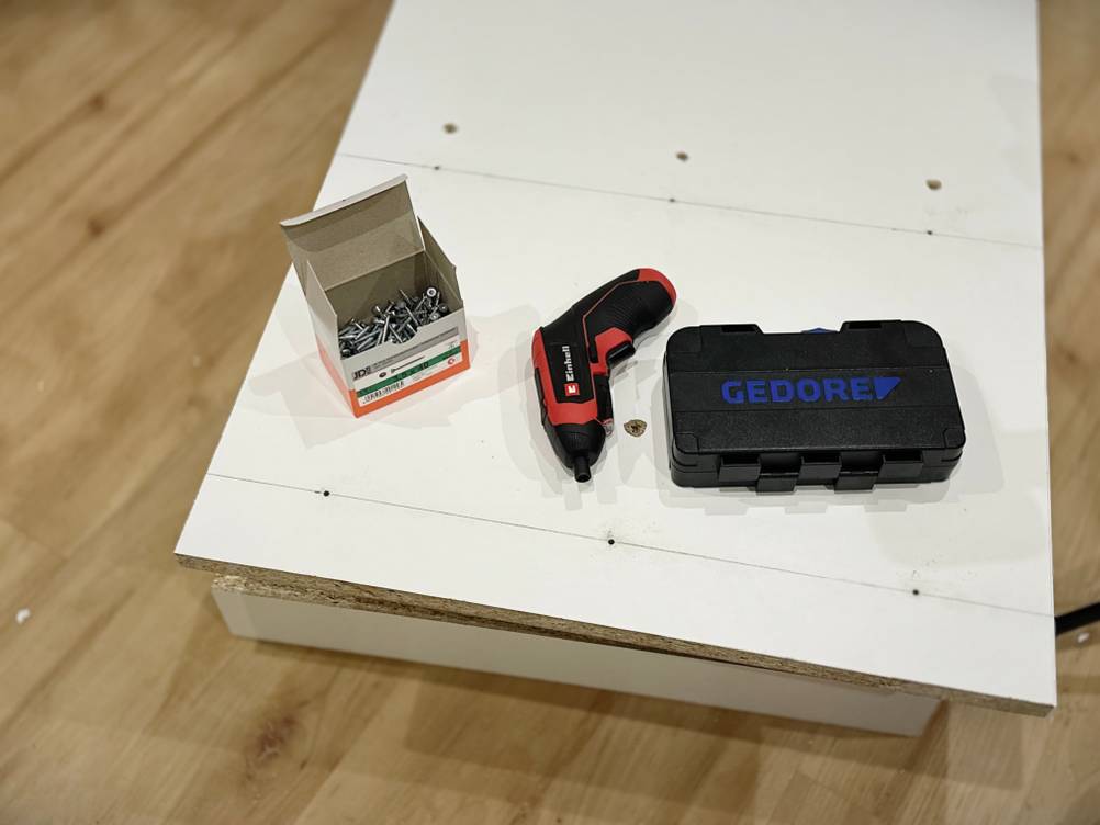 Ein Akkuschrauber, eine Schraubenschachtel und das Bitset von Gedore liegen auf einer weißen Spanplatte.