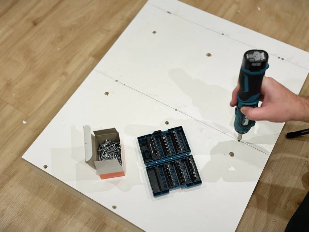 Das Bitset von Makita und eine Packung Schrauben liegen auf der weißen Spanplatte. Eine Hand hält einen Akkuschrauber.