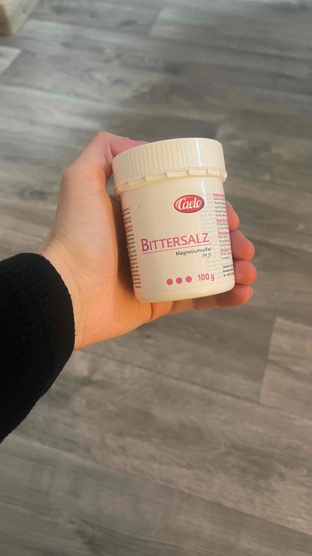 Eine Hand hält die weiße Schraubdose mit 100 Gramm des getesteten Bittersalzes über einem grauen Bodenbelag.