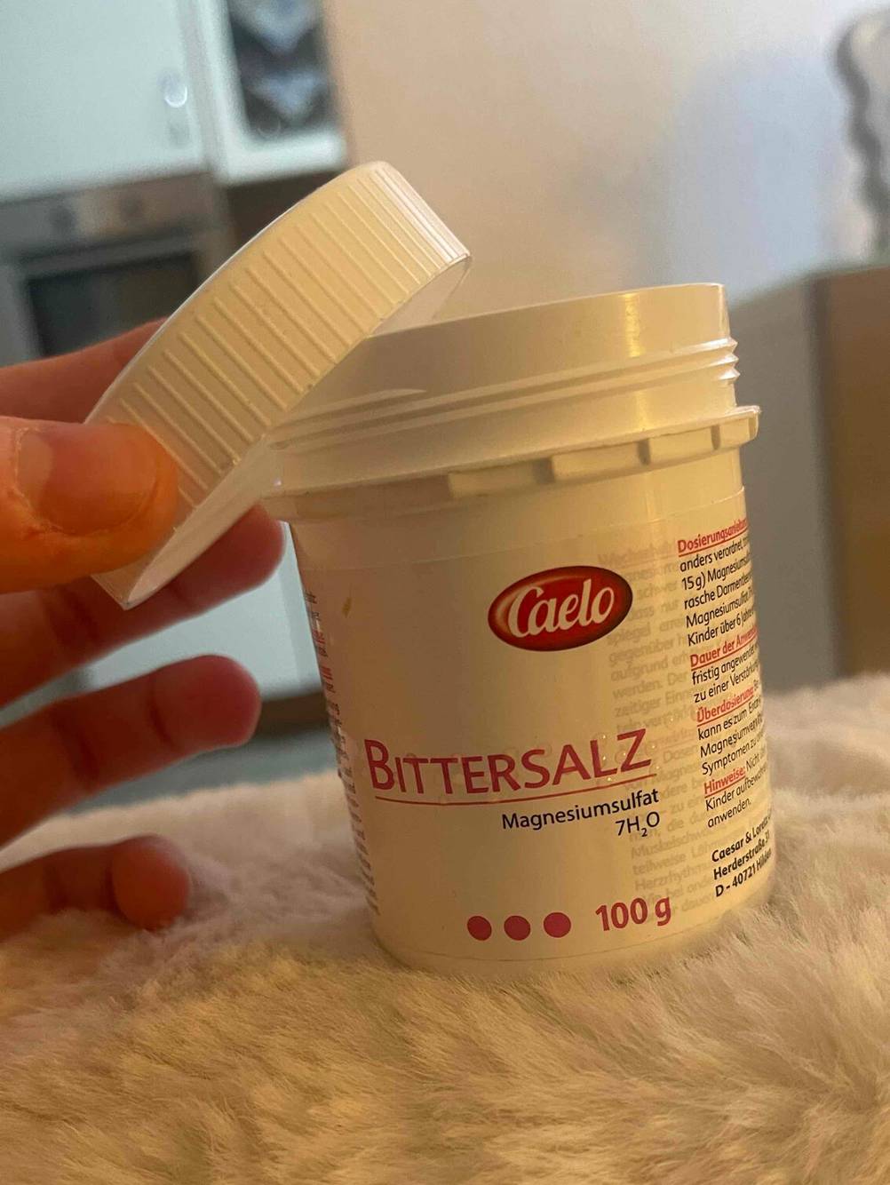 Eine Hand hält den abgenommenen Schraubverschluss über die weiße Dose mit 100 Gramm des getesteten Bittersalzes.